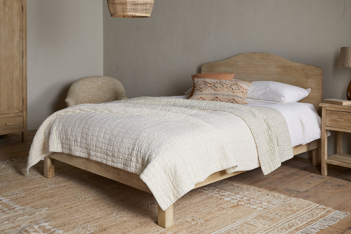 A cozy bedroom featuring the nkuku Vivan Grooved Bed in natural mango wood, complemented by a beige quilt, white sheets, decorative pillows, a matching wooden nightstand and wardrobe, woven chair, ceiling lamp, and a textured neutral rug on a wood floor.