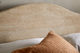 A close-up of the Vivan Grooved Bed - Natural by nkuku, featuring a solid wood headboard with horizontal grooves, a white bedsheet, and an orange-brown textured pillow. The light-colored wall behind has a subtly rough finish.