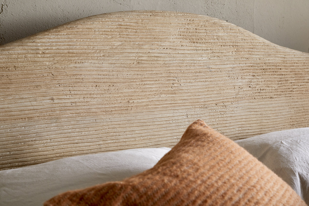 A close-up of the Vivan Grooved Bed - Natural by nkuku, featuring a solid wood headboard with horizontal grooves, a white bedsheet, and an orange-brown textured pillow. The light-colored wall behind has a subtly rough finish.