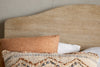 A close-up of the nkuku Vivan Grooved Bed - Natural’s textured wooden headboard behind two pillows—one rust-striped, the other beige with blue and tan embroidery—set against a light wall with visible cracks.