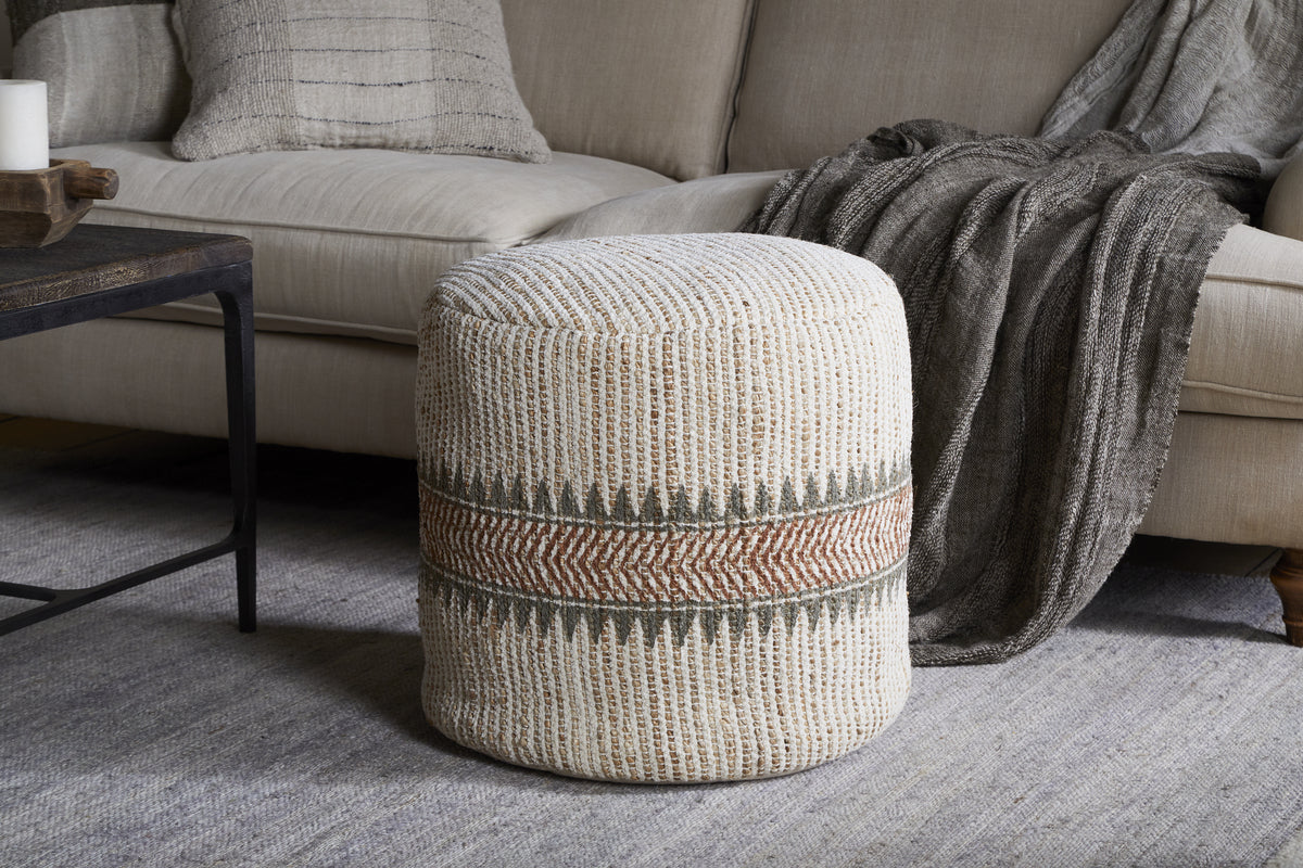 The nkuku Zairya Jute & Cotton Tall Pouf in natural and rust, featuring a textured cylindrical design with beige, green, and rust zigzag stripes, sits on a light gray rug before a beige sofa with gray accents and a dark wood coffee table.