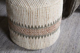 The nkuku Zairya Jute & Cotton Tall Pouf in Natural & Rust features textured, woven fabric with vertical beige and brown stripes and a central zigzag band of olive green and rust chevrons, shown on a gray woven rug.