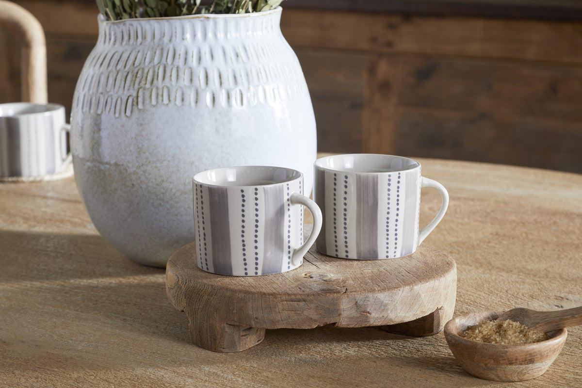 The nkuku Patthar Mug - Cream & Grey - Small (Set of 2) features hand-painted stoneware mugs with vertical dotted lines on a wooden stand. A white textured vase and a wooden spoon of brown sugar sit nearby on the table.