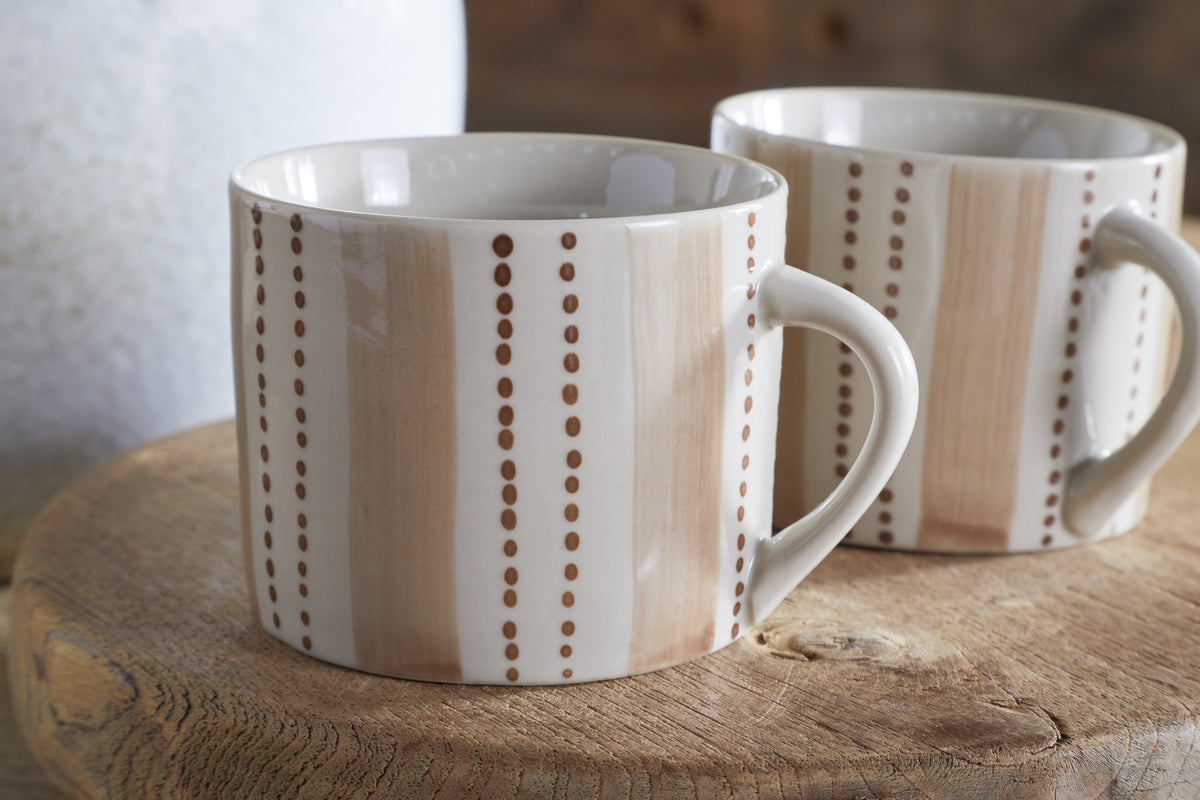 The nkuku Patthar Mug - Cream & Clay - Small (Set of 2) features hand-painted vertical beige stripes and brown dots, shown on a round wooden surface with a softly blurred background, part of the Patthar dinnerware collection.