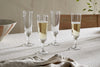 Four Pani Champagne Flutes by nkuku, in a set of 4 clear glasses, are arranged on a beige tablecloth; two are filled with bubbly champagne and two are empty. A folded napkin rests nearby alongside a striped bowl and leafy branch.