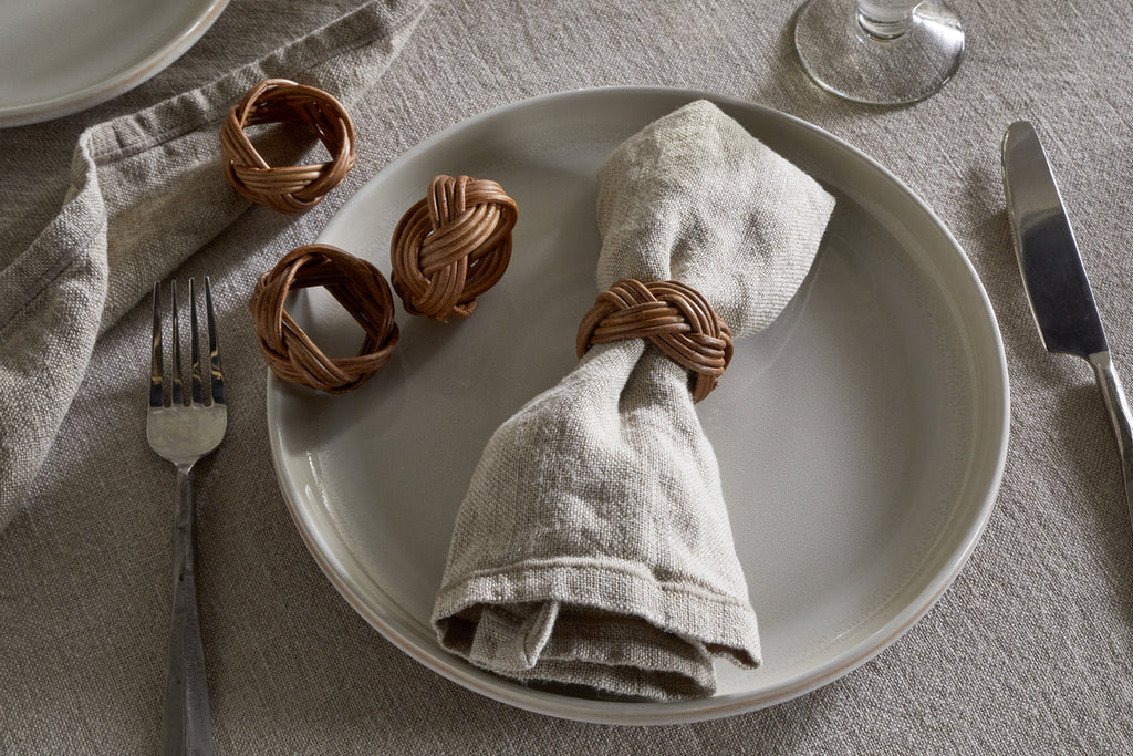 On a white plate sits a beige linen napkin with an nkuku Bersama Rattan Napkin Ring in Natural; two extra rings from the set of 4 rest nearby. Utensils and a glass complete the elegant look atop a neutral linen tablecloth.
