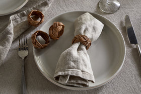 On a white plate sits a beige linen napkin with an nkuku Bersama Rattan Napkin Ring in Natural; two extra rings from the set of 4 rest nearby. Utensils and a glass complete the elegant look atop a neutral linen tablecloth.