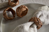 A close-up of three nkuku Bersama Rattan Napkin Rings – Natural – Set of 4, two on a plate and one holding a beige linen napkin, all on a neutral tablecloth; the rings showcase an intricate crisscrossed design for elegant table decor.