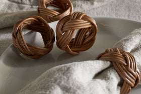 Close-up of nkuku's Bersama Rattan Napkin Rings—Natural, Set of 4—holding light beige linen napkins on a smooth, light gray plate. Another ring rests nearby, enhancing the inviting table decor against a soft, textured fabric background.