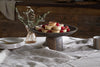 The nkuku Eisha Wooden Cake Stand in brown displays raspberry-topped tartlets on a linen-covered table, accompanied by elegant serveware: stacked plates, gold-colored forks, a striped mug, and a vase with olive branches.