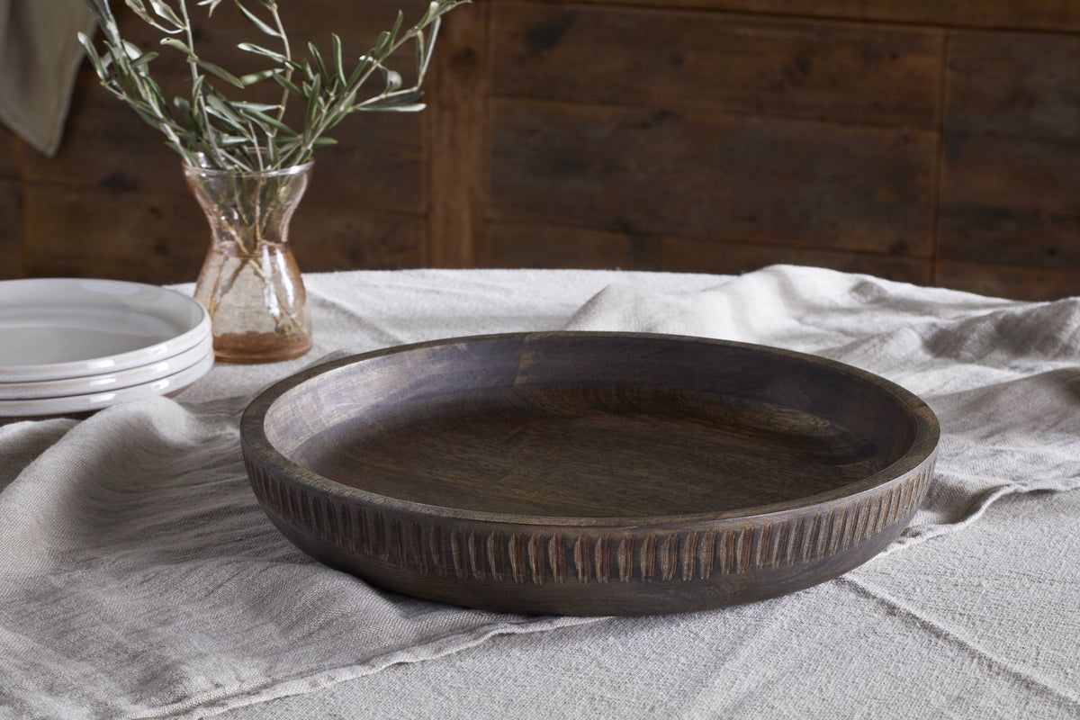The Eisha Bowl - Brown by nkuku, featuring a hand-carved rim, rests on a beige linen tablecloth. A small vase with greenery and stacked white plates complete the look against rustic wood paneling.