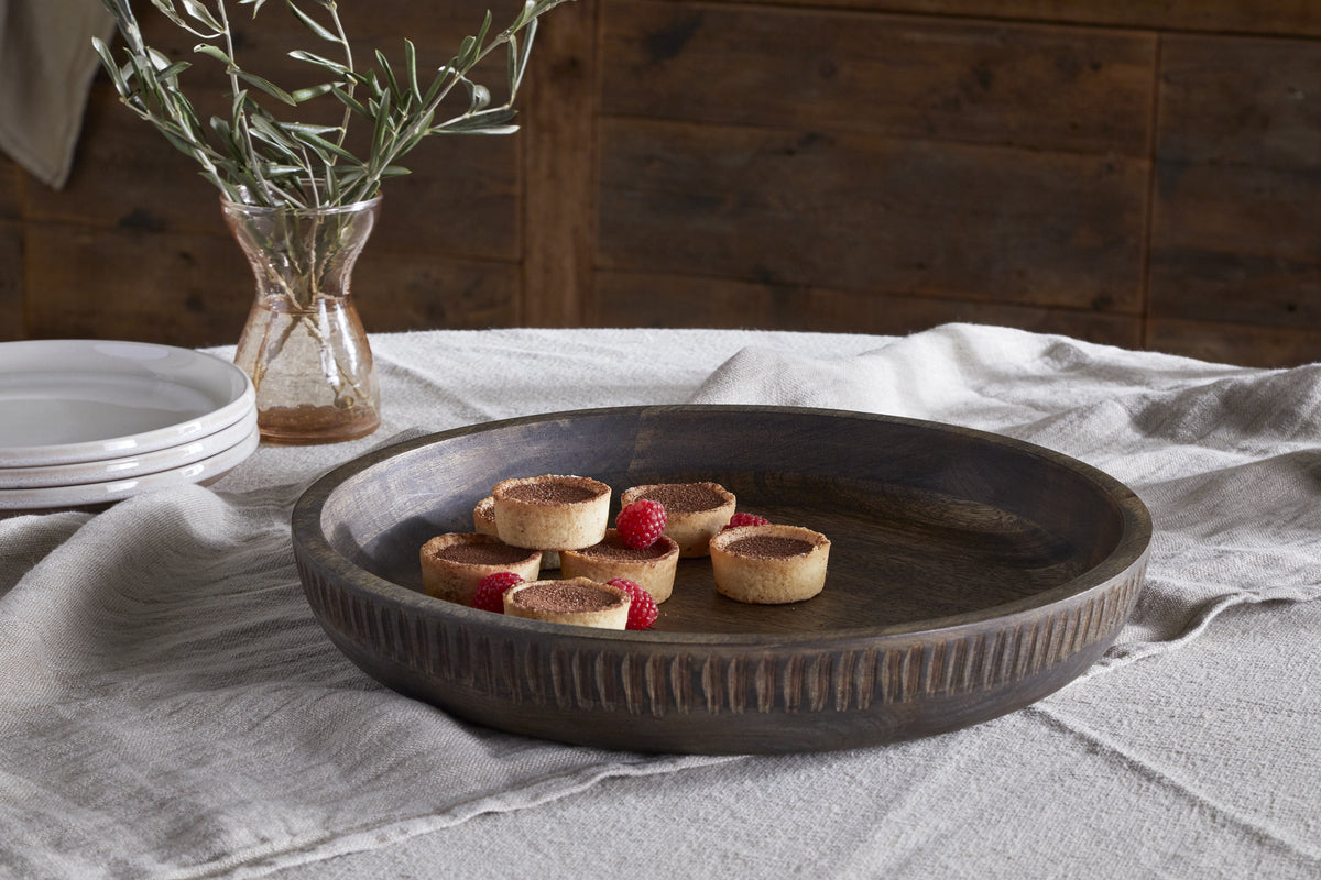 The Eisha Bowl - Brown by nkuku holds six small tarts, some topped with raspberries, on a textured white cloth. In the background, stacked white plates and a glass vase with green stems sit on a wooden table against a wooden wall.