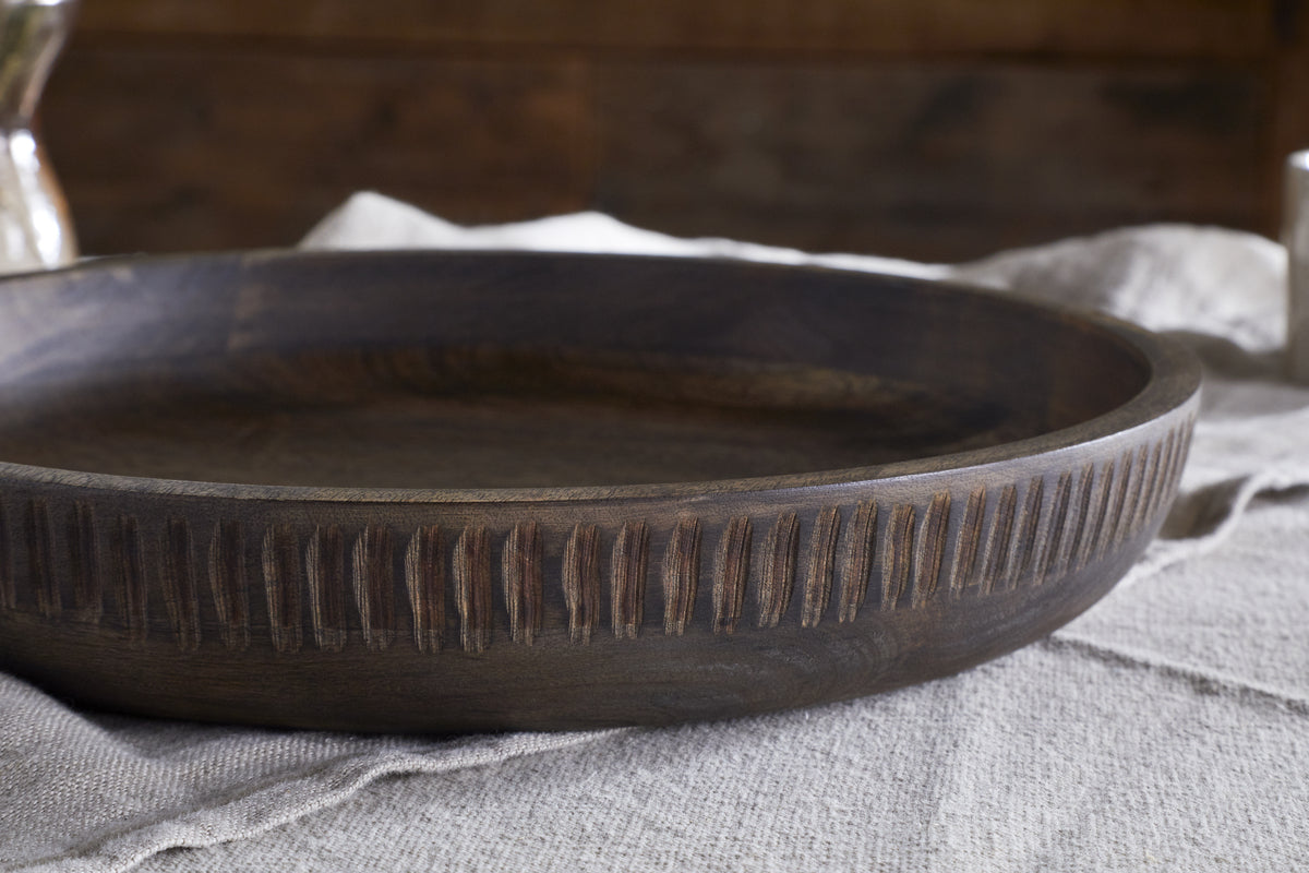 The Eisha Bowl - Brown by nkuku, featuring a round, hand-carved rim and vertical grooves, sits on light textured fabric with soft warm brown tones and a blurred ceramic object in the background.