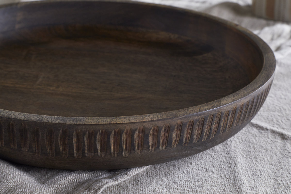 A close-up of the Eisha Bowl - Brown by nkuku shows its round, hand-carved rim with vertical grooves and a smooth, natural finish. The shallow wooden bowl rests on textured light fabric, with another object blurred softly in the background.