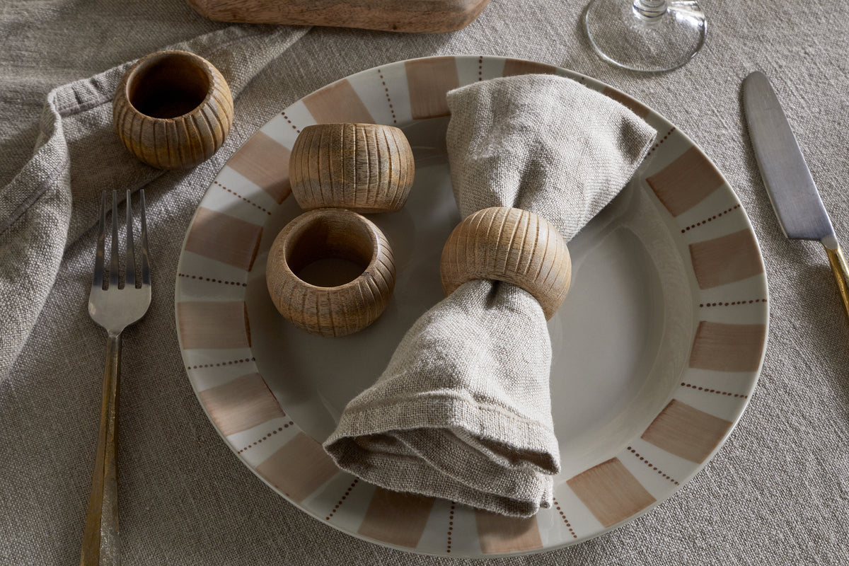 A beige linen napkin with an nkuku Vivan Napkin Ring (Natural, set of 4) rests on a patterned plate. Three extra rings are beside it, with cutlery and a wine glass atop the textured tablecloth for an elegant setting.