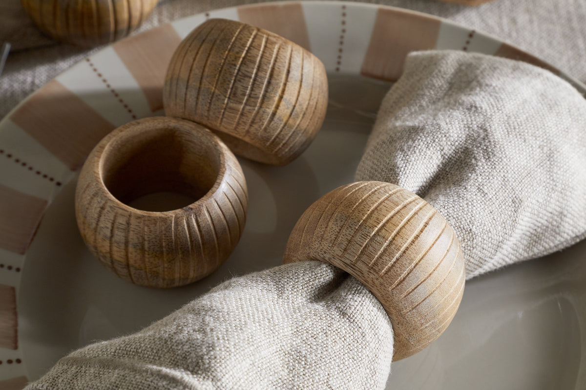 A close-up of nkuku’s Vivan Napkin Rings in Natural (set of 4) on a ceramic plate, with one ring holding an off-white linen napkin and the others arranged nearby, all elegantly displayed in soft natural light.