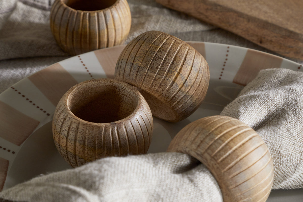 Close-up of nkuku's Vivan Napkin Rings in Natural (Set of 4), featuring carved vertical lines and holding beige linen napkins on a patterned plate. An extra ring sits nearby, enhancing the table decor with rustic charm.