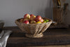 The Bersama Rattan Fruit Bowl by nkuku, filled with apples and a lime, sits on a rustic wooden table—a charming kitchen centerpiece with utensils in a wicker holder and a folded cloth in the background.