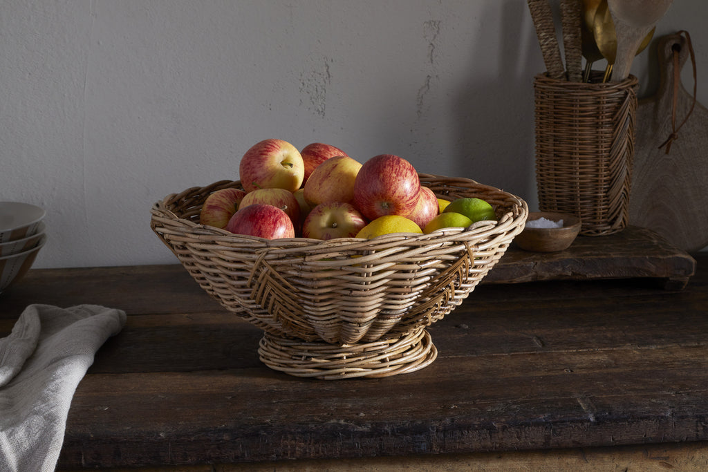 The Bersama Rattan Fruit Bowl by nkuku, filled with apples and a lime, sits on a rustic wooden table—a charming kitchen centerpiece with utensils in a wicker holder and a folded cloth in the background.