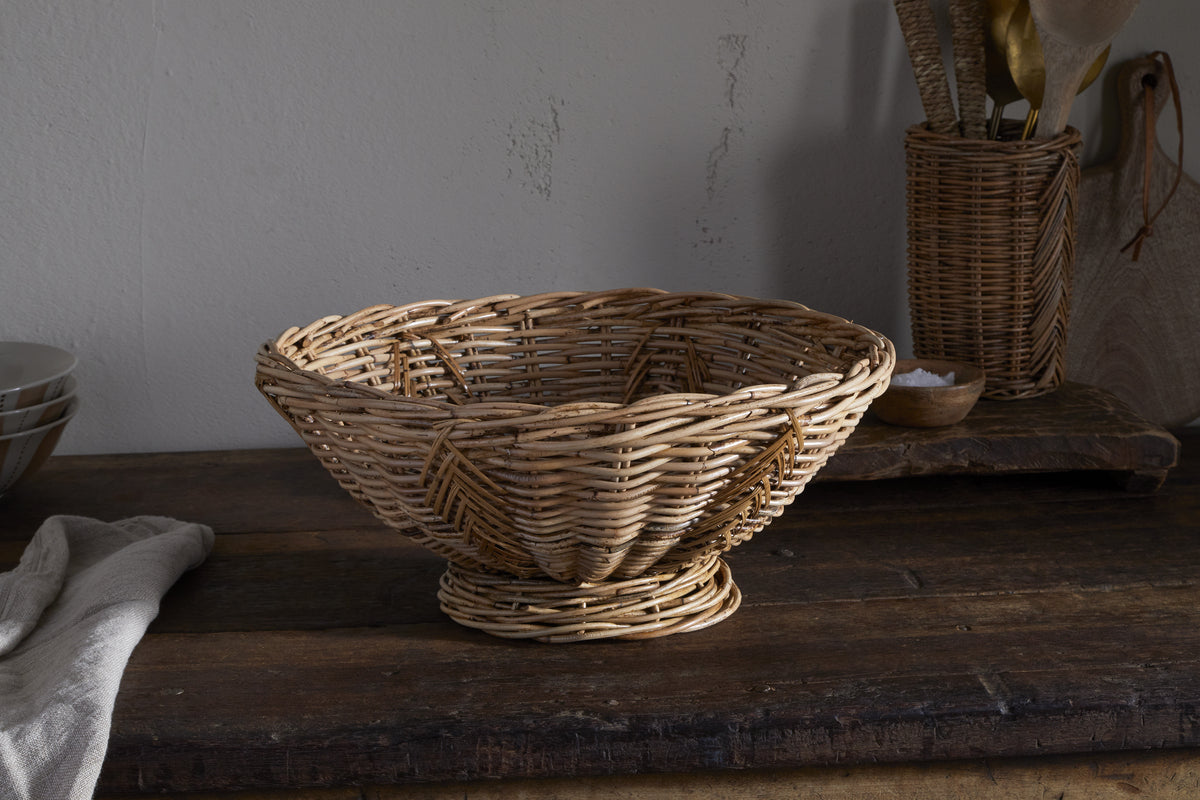 The Bersama Rattan Fruit Bowl by nkuku, with its wide, shallow shape, sits on a rustic table alongside a white towel, stacked bowls, and a wicker utensil holder—soft natural light enhances this inviting kitchen centrepiece.