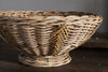 The Bersama Rattan Fruit Bowl by nkuku features a round, handwoven design with a wide rim and narrow base in natural tan. Set on a dark wooden surface against a textured wall, it adds rustic charm as a kitchen centrepiece.