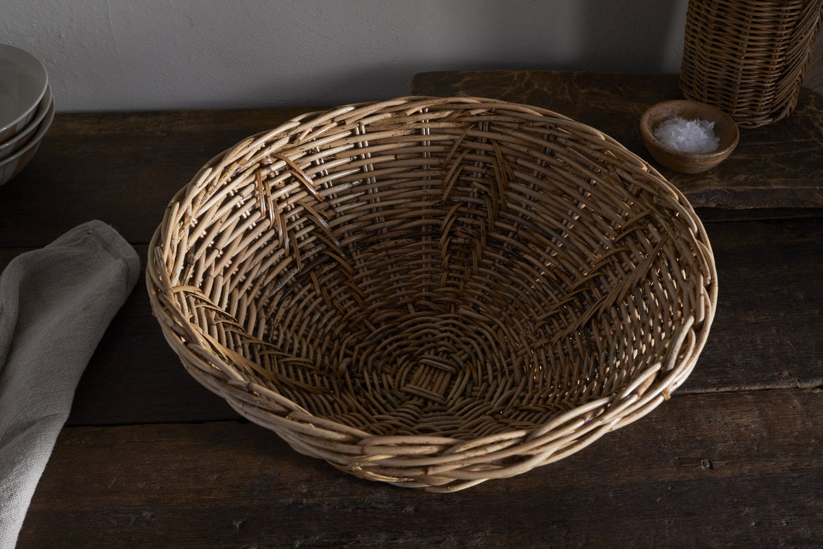 The Bersama Rattan Fruit Bowl - Natural by nkuku, large and handwoven, rests empty on a dark wooden surface with a folded beige cloth to the left and white plates stacked in the background. A small bowl of salt sits nearby.