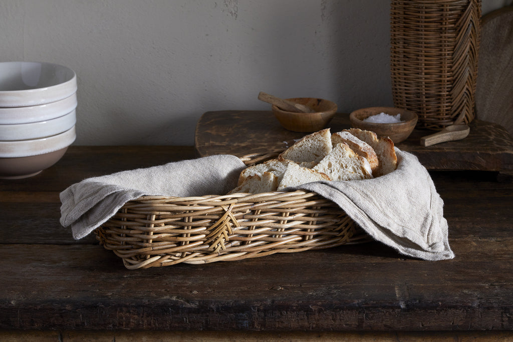 The nkuku Bersama Rattan Bread Basket - Natural, lined with beige cloth, displays rustic bread on a wooden table. Stacked white bowls and wooden salt bowls complete the cozy, rustic kitchen decor in the background.