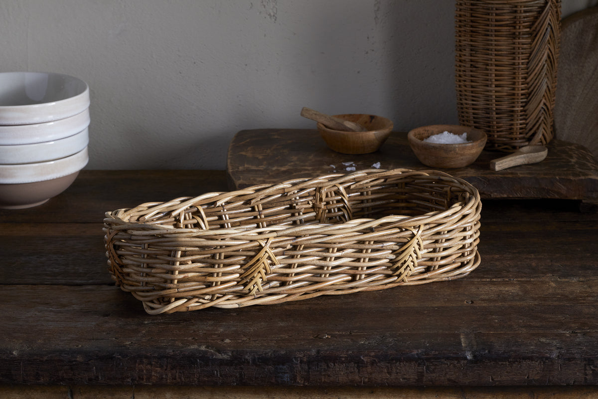 The Bersama Rattan Bread Basket by nkuku sits on a rustic wooden table, with stacked white bowls, small wooden salt bowls, a scoop, and a tall wicker container creating charming decor against a light wall.