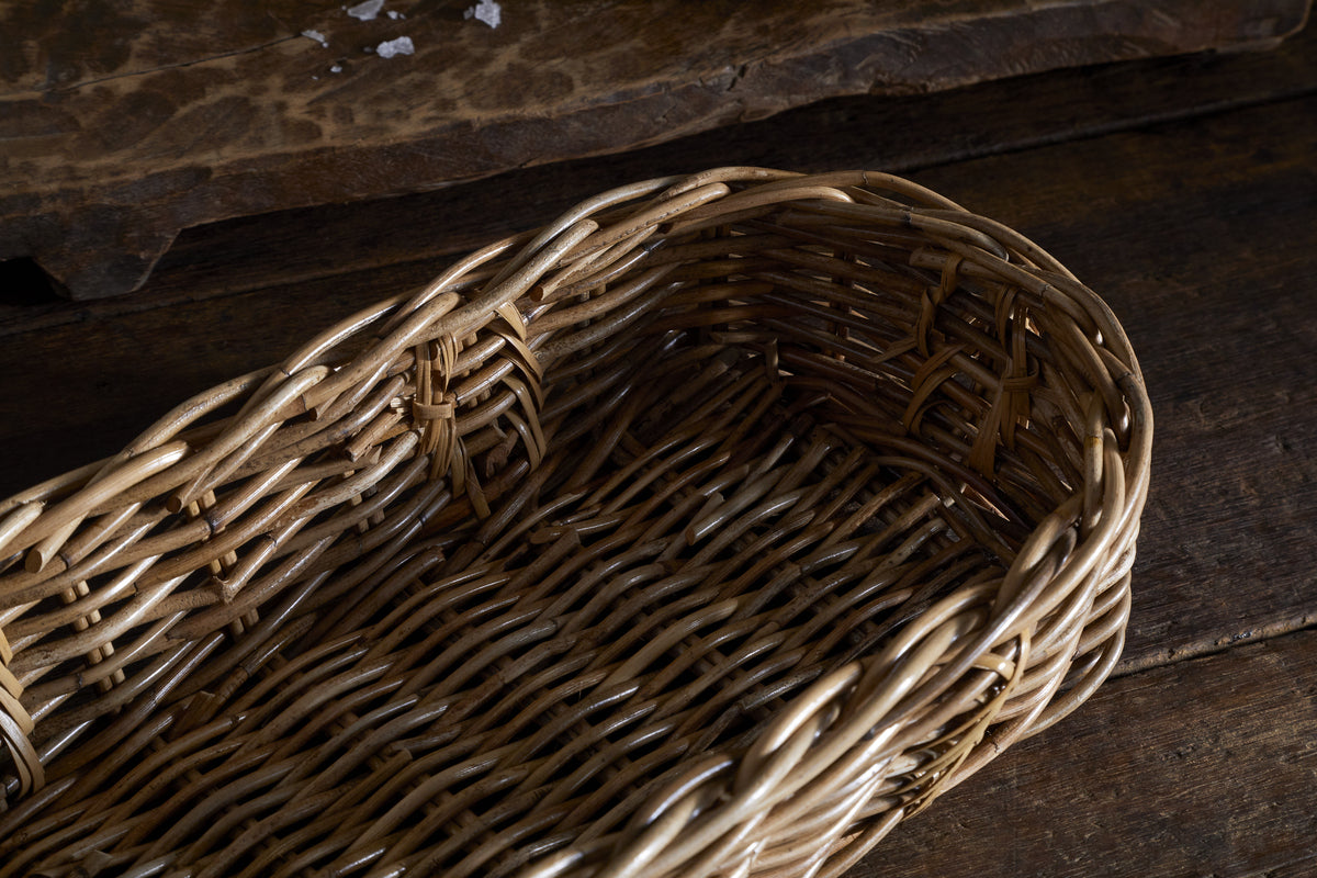 The Bersama Rattan Bread Basket by nkuku is shown close-up, empty and angled to display its handwoven pattern and side rim, resting on a rustic wooden surface—ideal as tabletop decor under warm lighting.