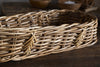 The Bersama Rattan Bread Basket - Natural by nkuku is an empty, oval handwoven basket made from light and dark brown rattan reeds. It sits on a rustic wooden surface with blurred wooden bowls adding charm to the warm tabletop decor.