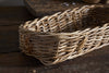 The Bersama Rattan Bread Basket by nkuku features a tight, handwoven rectangular design in natural rattan. It rests on rustic wood, making it ideal for tabletop décor while lighting accentuates its textured wicker and wooden details.