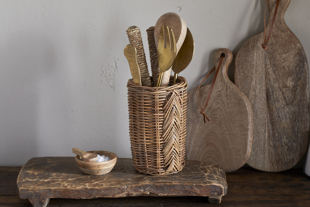 The nkuku Bersama Rattan Utensil Holder - Natural, filled with assorted wooden and brass cutlery, rests on a rustic surface next to a small wooden salt bowl. Wooden cutting boards hang on the wall in the background.