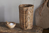 The Bersama Rattan Utensil Holder by nkuku, a tall cylindrical handwoven basket with a chevron pattern, sits on a rustic wooden surface—ideal for kitchen storage. Nearby are a small wooden bowl with salt and scoop, and a cutting board in the background.