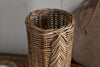 The nkuku Bersama Rattan Utensil Holder - Natural is a cylindrical, handwoven basket with a chevron pattern and natural brown color, displayed on a wooden surface against a neutral textured wall.