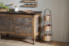 A rustic kitchen showcases a wooden cabinet, jars, a plant, and wall shelf. Next to it stands the nkuku Montali Iron & Rattan Three-Tiered Storage Stand in Natural, neatly holding baskets of assorted fruits and vegetables.