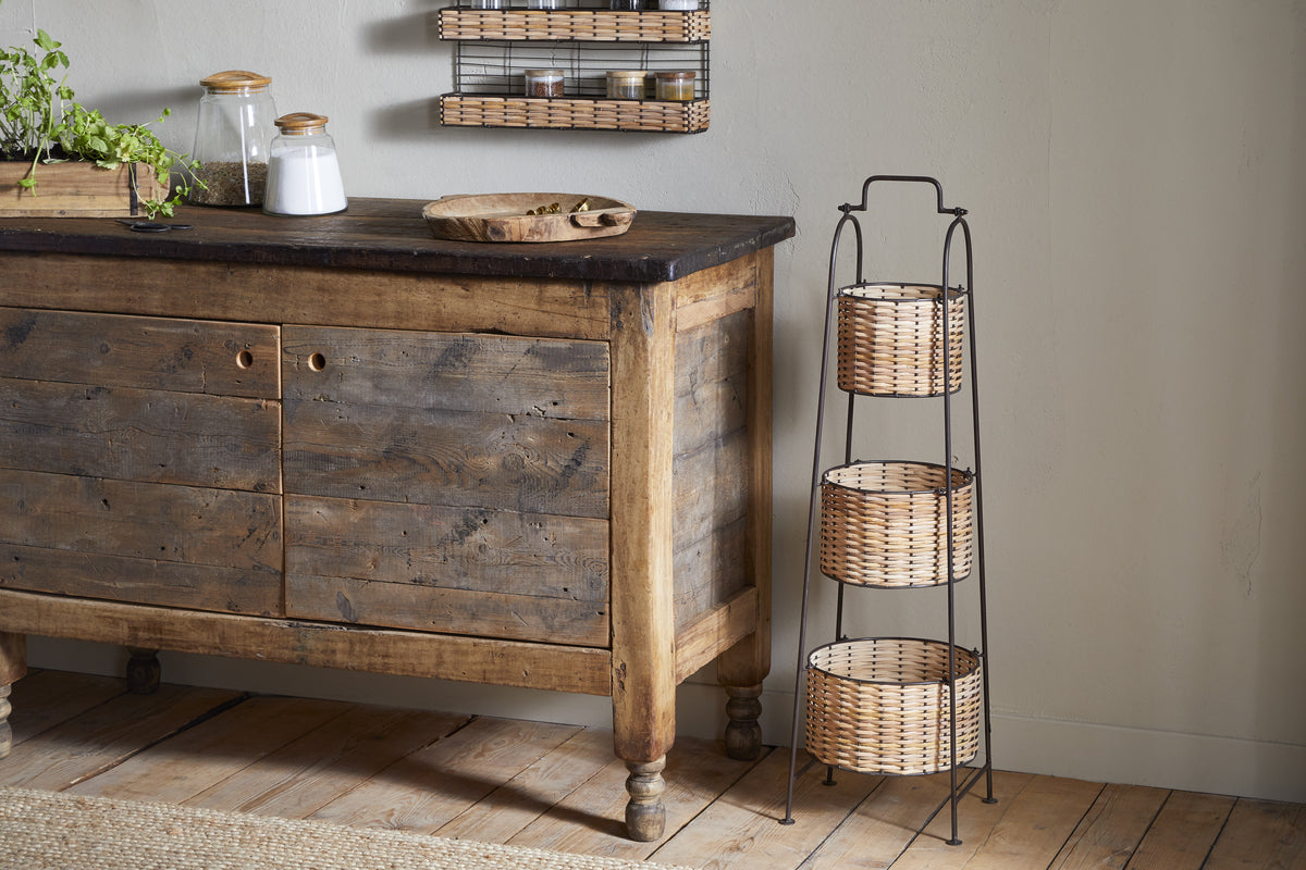 Against a light wall, jars and a wooden tray sit on a rustic sideboard. Next to it stands the nkuku Montali Iron & Rattan Three-Tiered Storage Stand—ideal for storing kitchen or pantry essentials in its handwoven baskets. The floor is wood.