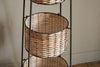 The nkuku Montali Iron & Rattan Three-Tiered Storage Stand - Natural features round handwoven baskets on a dark metal frame, ideal for kitchen or pantry organization. Shown on a wooden floor against a textured beige wall.