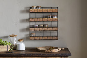 A rustic kitchen scene features a wooden table with jars, spoon, and herbs, while the nkuku Montali Iron & Rattan Spice Rack - Natural adds stylish storage with handwoven rattan on the light-colored wall.