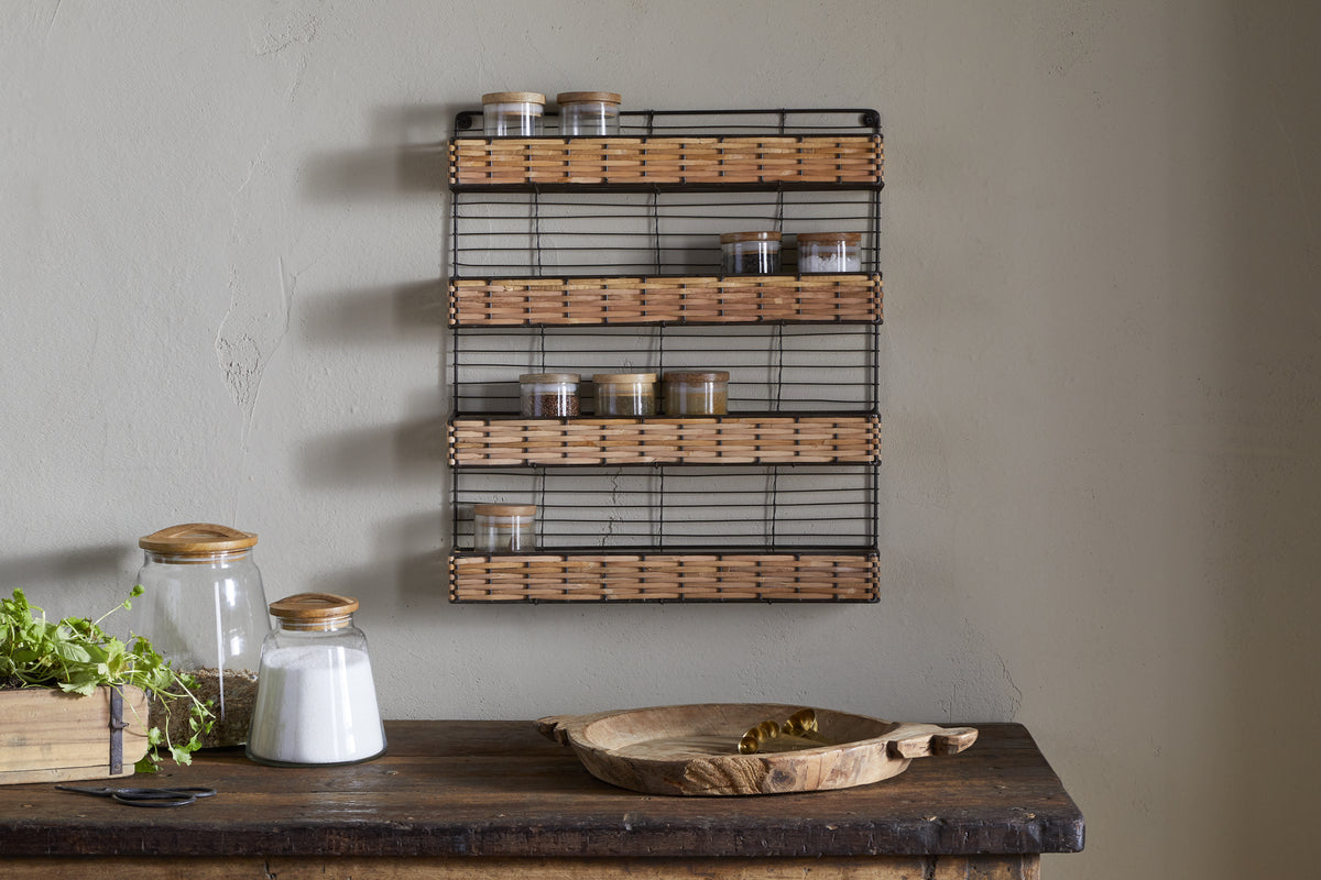 A rustic kitchen scene features a wooden table with jars, spoon, and herbs, while the nkuku Montali Iron & Rattan Spice Rack - Natural adds stylish storage with handwoven rattan on the light-colored wall.