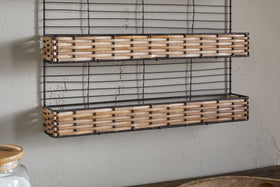 The nkuku Montali Iron & Rattan Spice Rack - Natural features two handwoven rattan shelves with a black wire frame. Wall-mounted above a wooden surface on a light gray wall, it's ideal for kitchen storage. A glass jar with a wooden lid sits nearby.