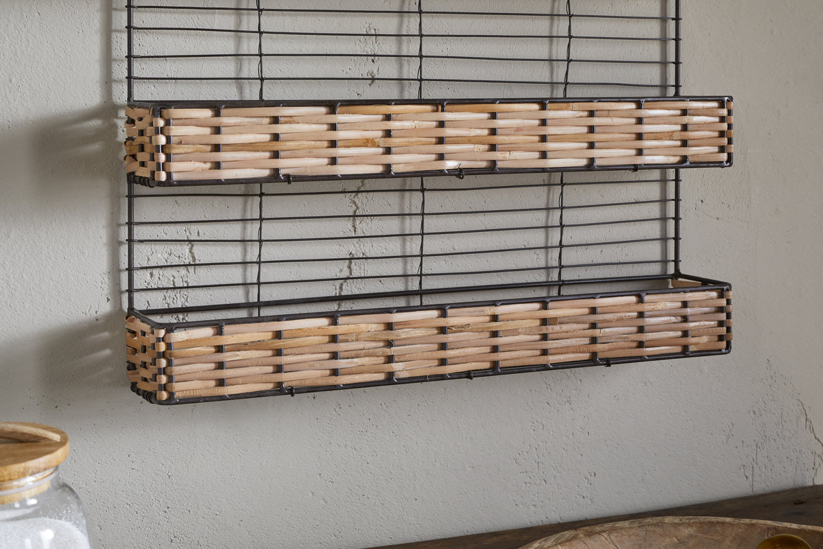 The nkuku Montali Iron & Rattan Spice Rack - Natural features two handwoven rattan shelves with a black wire frame. Wall-mounted above a wooden surface on a light gray wall, it's ideal for kitchen storage. A glass jar with a wooden lid sits nearby.