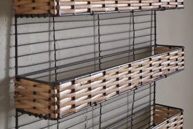 Close-up of the nkuku Montali Iron & Rattan Spice Rack - Natural, wall-mounted on a light grey textured wall. The rack has three empty rectangular wire baskets with handwoven rattan trim and black metal wire sides.