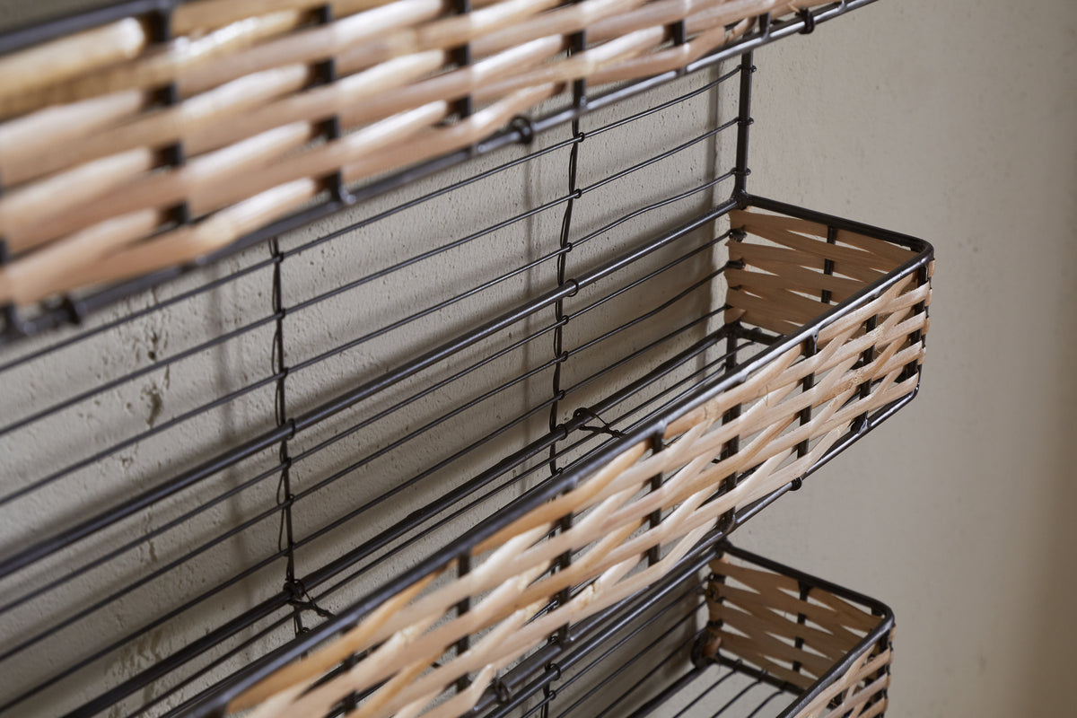 Close-up of nkuku's Montali Iron & Rattan Spice Rack - Natural, a wall-mounted organizer with three wire baskets and handwoven rattan fronts. The black frame contrasts with the light wall—ideal for kitchen storage or spices.