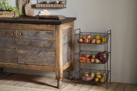 A rustic wooden cabinet sits on a wood floor beside the nkuku Inkollu Three Tier Storage Stand in antiqued brass, filled with apples, carrots, onions, and potatoes. A loaf of bread is on a cutting board atop the cabinet; kitchen items and greenery are visible.