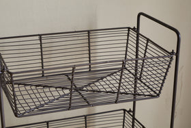 A close-up of the nkuku Inkollu Three Tier Storage Stand in antiqued brass, ideal for kitchen storage. The top rectangular basket displays its crisscross wire design, with another empty basket partially visible beneath.
