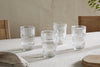 A set of four nkuku Kaanch Highball glasses in clear glass, featuring horizontal ridges, are arranged on a beige tablecloth with wooden chairs and a cutting board softly visible in the background.