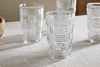 The nkuku Kaanch Highball - Clear (Set of 4) features textured, geometric glassware on a light woven fabric surface—three grouped in the back and one separately displayed in brighter light at the front.