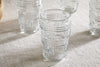 The nkuku Kaanch Highball - Clear (Set of 4) features embossed geometric glass tumblers, displayed on a light beige fabric with one centered in focus and three softly blurred behind.