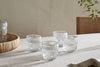 Four nkuku Kaanch Tumblers - Clear (Set of 4) with an embossed check pattern rest on a light tablecloth. A wooden tray with another Kaanch tumbler is on the left, and a chair with green stems appears to the right against a smooth wall.