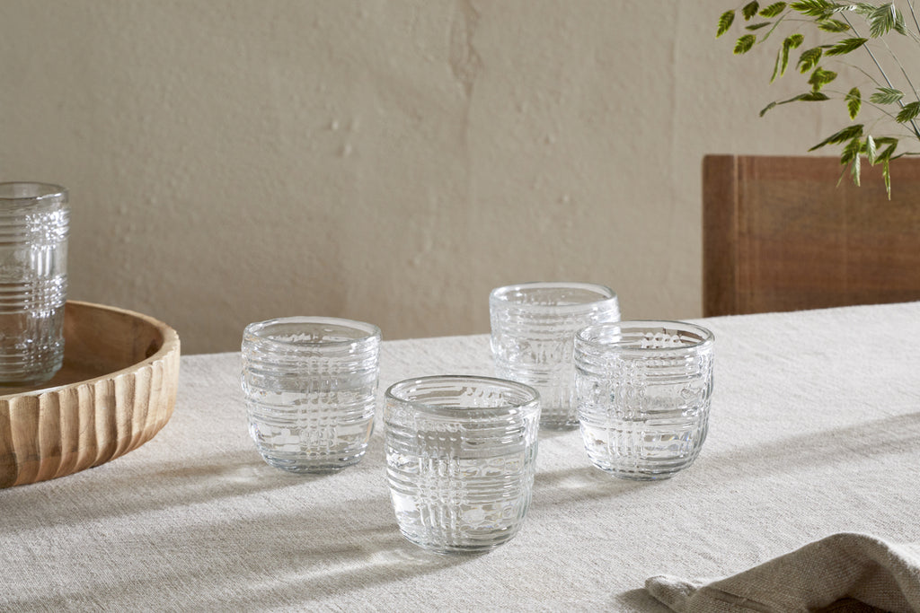 Four nkuku Kaanch Tumblers - Clear (Set of 4) with an embossed check pattern rest on a light tablecloth. A wooden tray with another Kaanch tumbler is on the left, and a chair with green stems appears to the right against a smooth wall.